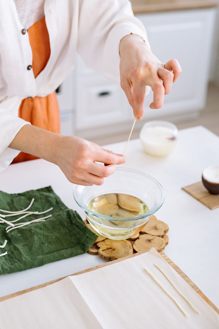 Person making candles by dipping wicks into wax in a home kitchen setting.