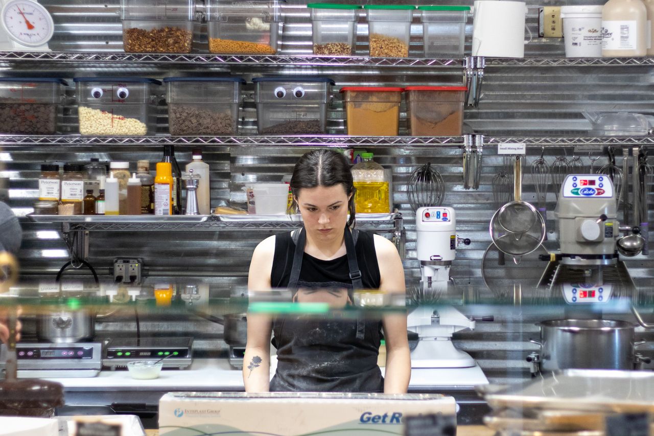 Woman working in a commercial kitchen surrounded by spices and machinery, focusing on her task.